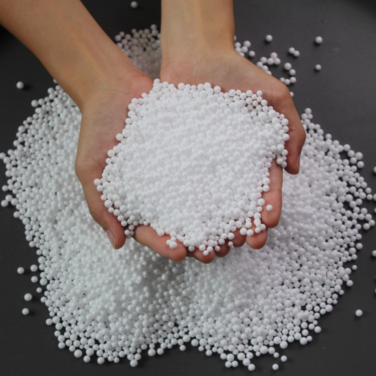 Hands holding a pile of white expanded polystyrene beads, with additional beads scattered on a dark surface.